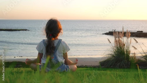 A girl sits in a meditation pose on the beach at sunset. A girl doing yoga on the beach. A young girl watching the ocean at the green field in 4K. enjoying of the calm of the evening sunset sea. High