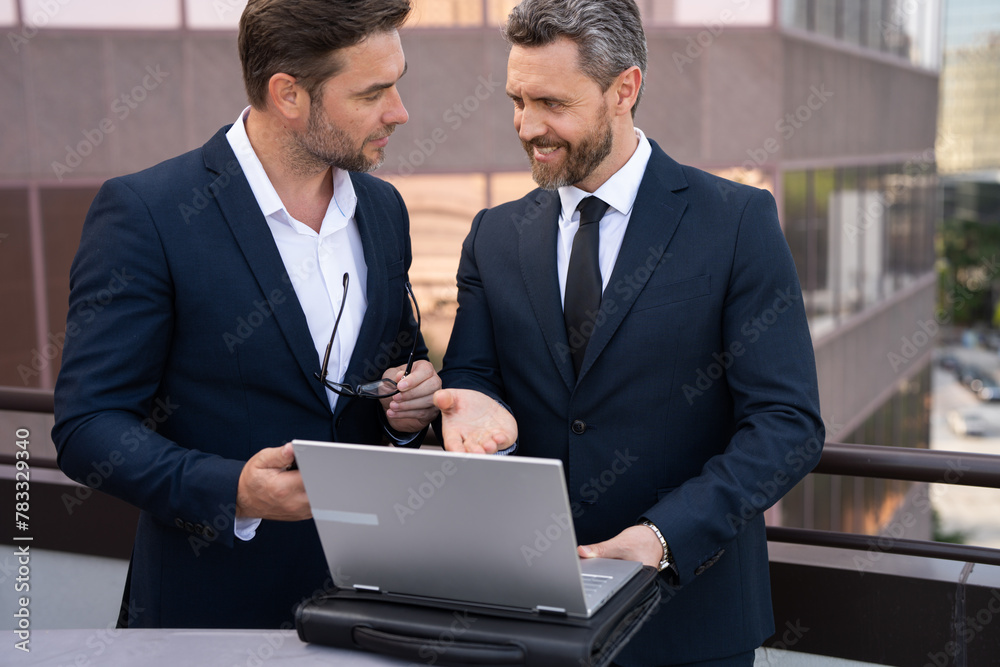 Two businessmen on the street, deeply engrossed, eyes fixed on their laptop screen, perhaps discussing a crucial deal or strategizing for their next big project in the bustling cityscape. Two businessmen on the street, deeply engrossed, eyes fixed on their laptop screen, perhaps discussing a crucial deal or strategizing for their next big project in the bustling cityscape.