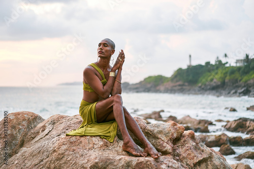 Gay bipoc graceful man poses sitting high on rocks in ocean at sunset. Homosexual slim ethnic fashion model in tropical maxi dress on top of rocky hill above storm at dusk. Pride LGBTQIA