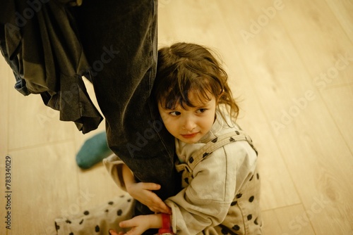 portrait of a small, capricious 3-year-old girl who clings to her mother's legs with her hands and feet and does not let her go, constantly asking for something and crying