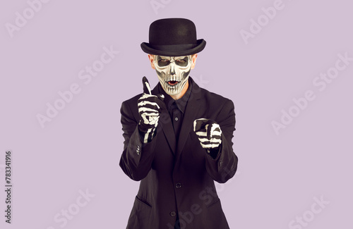 Aggressive Horror Movie Hero shoots at viewer with imaginary guns. Image of unkillable, ever-living mafia. Man in dark suit and black and white make-up. Isolate on light purple background.