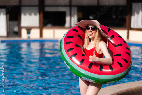 Beautiful girl in a red swimsuit, hat and sunglasses poses with an inflatable circle in the shape of a watermelon near the pool. Loves to relax in the pool on a sunny day. Shows thumbs up