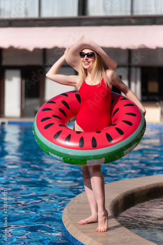 Beautiful girl in a red swimsuit, hat and sunglasses poses with an inflatable circle in the shape of a watermelon near the pool. Likes to relax in the pool on a sunny day