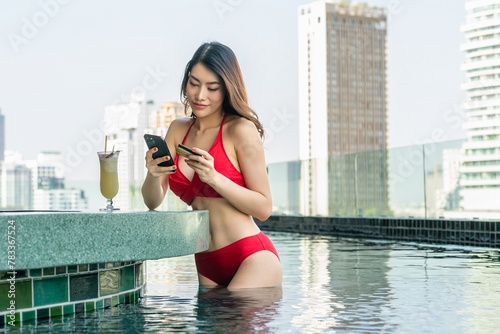 Poolside Banking: Young Asian woman in a red bikini conducts online banking on her phone while holding a credit card at the poolside bar counter