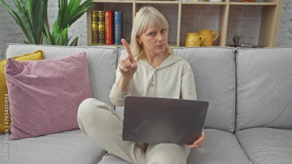 A middle-aged blonde woman gestures disapproval while using a laptop on a grey sofa indoors. A middle-aged blonde woman gestures disapproval while using a laptop on a grey sofa indoors.