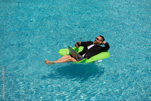 Funny business man in a business suit works for a laptop standing in the water in pool. Remote work. Crazy freelancer. Business and summer. Business man working online with computer laptop in pool.