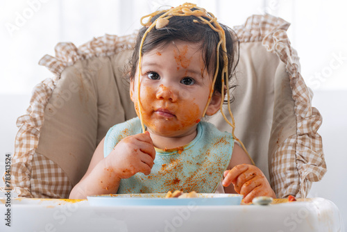 Adorable messy little child girl hungry use hand eating spaghetti sitting in high-powered chair at home. Toddler child with tomato sauce making mess face looking at food. Self-feeding concept