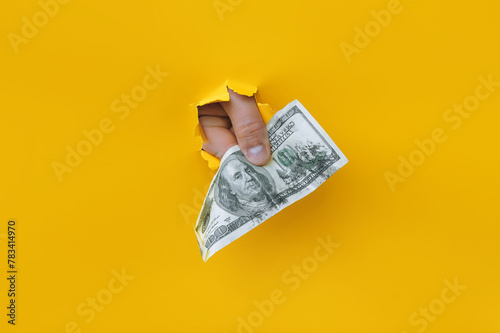 A right man's hand holds dirty money through a torn hole in yellow paper. Concept of dishonest income, donation, profit and salary fraud.
