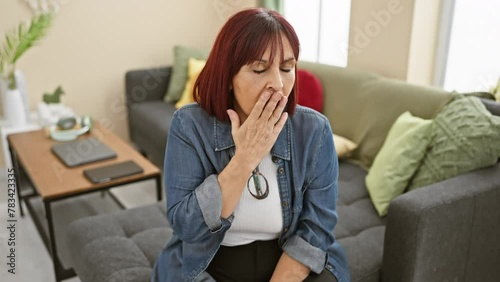 Tired and bored middle-aged hispanic woman, sleepily yawning with hand over mouth in a shirt at home photo