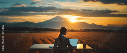 A panoramic, widescreen photo of a brown female office worker sitting at a desk overlooking a mountain range and a field at sunrise -  a laptop and a cup of coffee sit on the desk; working remotely photo