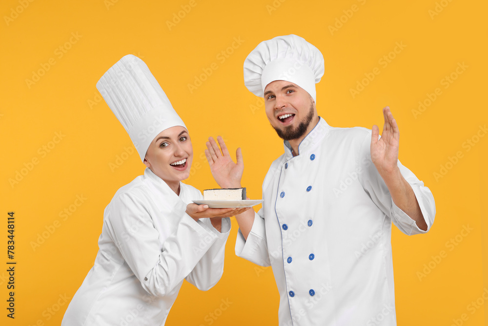 Happy professional confectioners in uniforms holding delicious cheesecake on yellow background