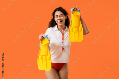 Beautiful young happy African-American female lifeguard with flippers on orange background