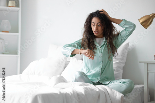 Overslept young African-American woman looking at wristwatch in bedroom
