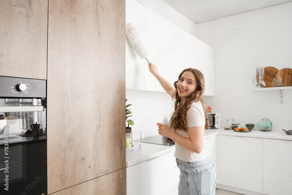 Beautiful young woman with pp-duster cleaning cupboard in kitchen