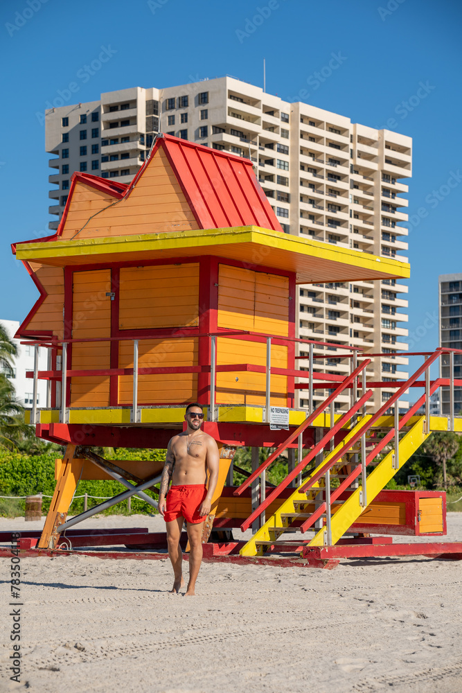 Young male tourist with a well-groomed body, having his picture taken at one of the lifeguard booths in Miami beach, on a beautiful sunny day.