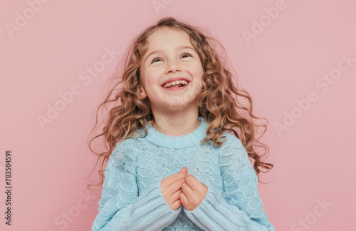 A cute little girl with long curly hair, wearing sweater is smiling and holding her hands on chest against pink background,