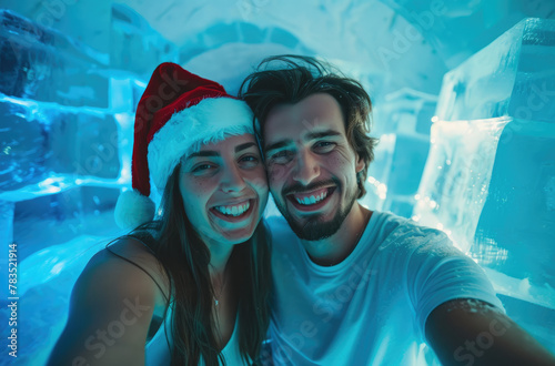 Photo of a happy couple taking a selfie with the front camera, wearing white t-shirts and Christmas hats inside an ice house with blue decorations © Kien