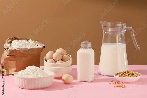 Front view of cooking or baking ingredients includes fresh milk in unlabeled glass bottle, eggs and flour in white round bowl, soybean in a small bowl with some seed lay flat on table. 