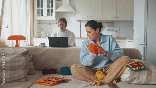 Young woman knitting crocheting with colored yarn granny square while watching online tutorial on phone. And young man working on laptop from homeWoman watching needlework lessons, home hobbies photo