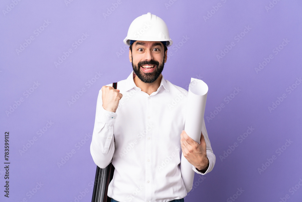 Young architect man with helmet and holding blueprints over isolated purple background celebrating a victory in winner position