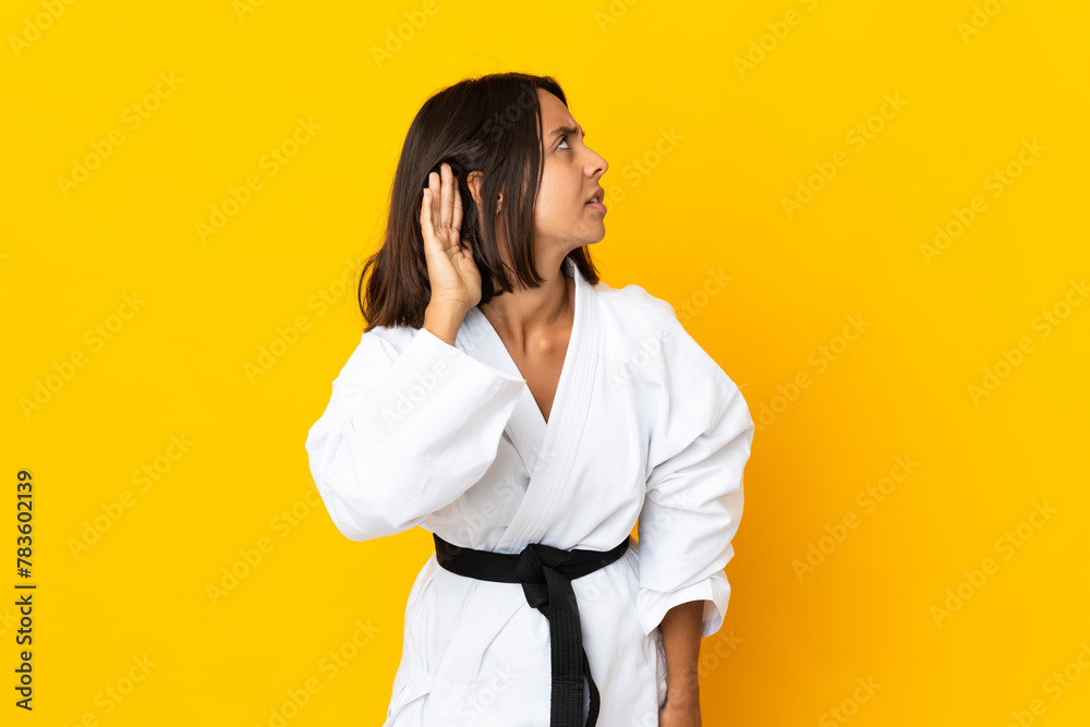 Young woman doing karate isolated on yellow background listening to something by putting hand on the ear Young woman doing karate isolated on yellow background listening to something by putting hand on the ear