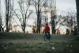 A relaxed young man cycling through a serene park with bare trees and a setting sun in the background.
