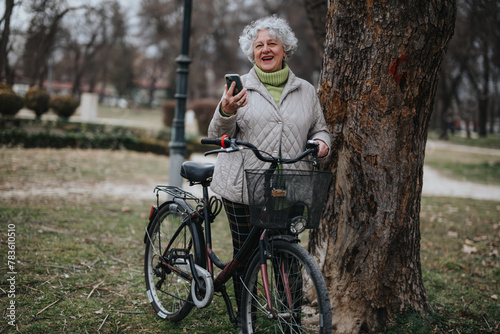 Joyful senior woman with curly grey hair standing by her bicycle in the park, holding a smart phone and smiling.