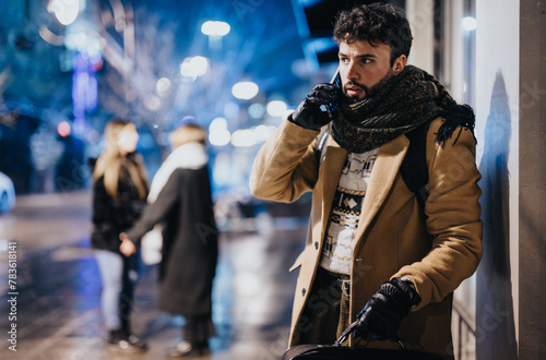 Focused young adult in warm clothes using a smart phone outdoors at night, with blurred city life and pedestrians in the backdrop.