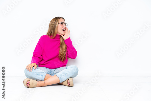 Young caucasian woman sitting on the floor isolated on white background shouting with mouth wide open to the lateral