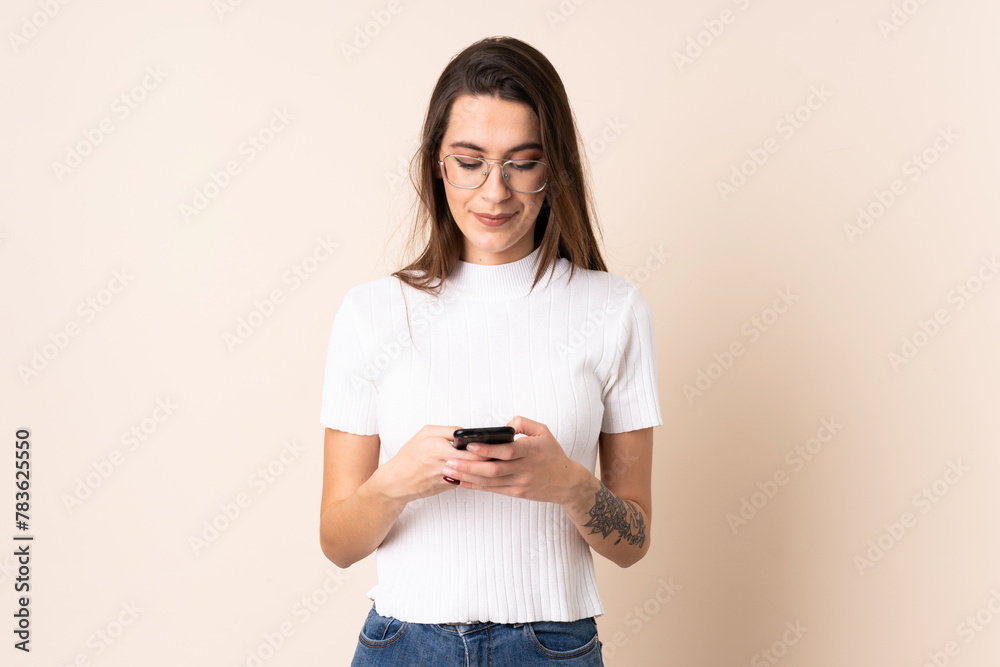 Young woman over isolated beige background sending a message with the mobile Young woman over isolated beige background sending a message with the mobile