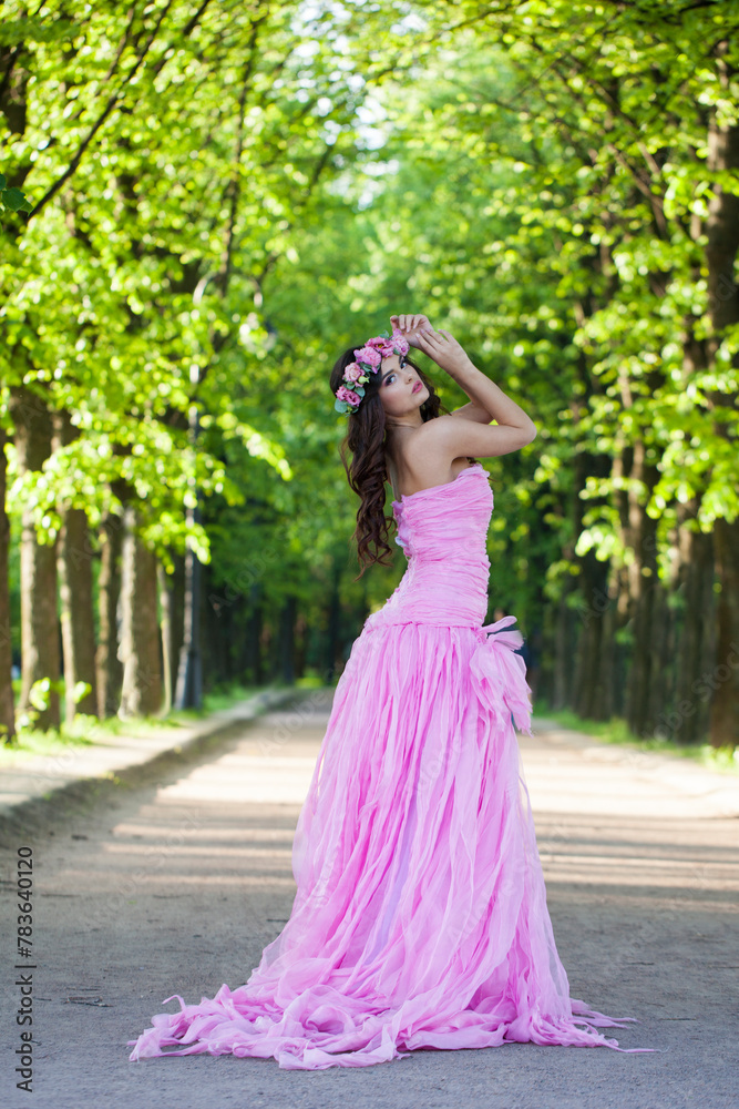 Glorious female model with bright makeup, curly hair and pink flower wreath on green summer foliage background. Healthy woman in pink dress portrait Glorious female model with bright makeup, curly hair and pink flower wreath on green summer foliage background. Healthy woman in pink dress portrait