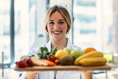 Beautiful smiling nutritionist holding wooden table with fresh fruits and vegetables in a medical consultation