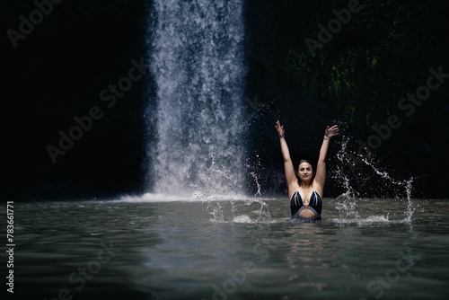 Caucasian woman swimming in pond and looking at camera with hands up in background of waterfall
