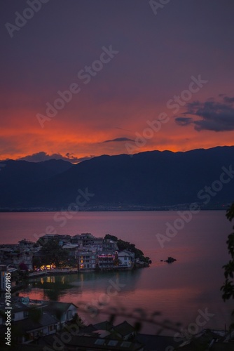 Vertical shot of coastal buildings separated by waters with hills and orange sunset in background