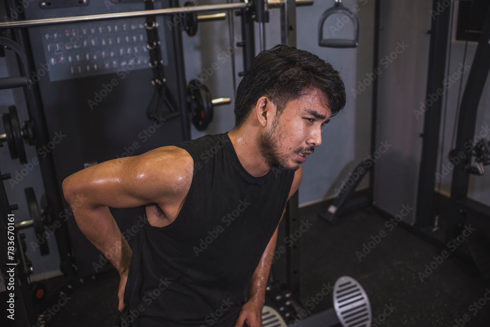 Naklejka premium A young asian man suffers from lower back pain while working out at the gym. Poor posture or overuse leading to chronic injury.