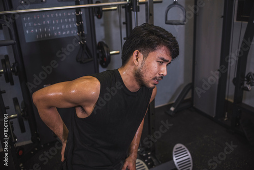 A young asian man suffers from lower back pain while working out at the gym. Poor posture or overuse leading to chronic injury.
