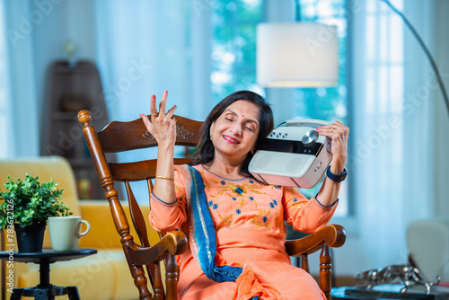 Indian senior adult woman having fun listening to radio at home while sitting on rocking chair.