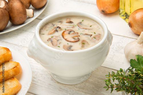 Cream of mushroom soup in a white porcelain tureen on a white rustic table with ingredients. Selected focus.
