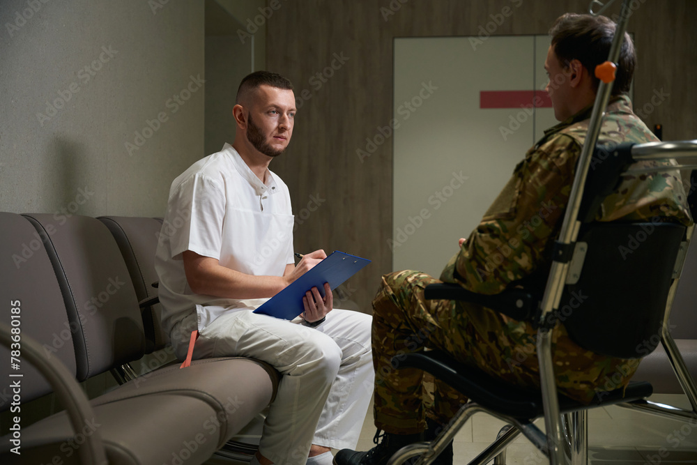 Physician writing down military patient complaints in hospital corridor - obrazy, fototapety, plakaty 