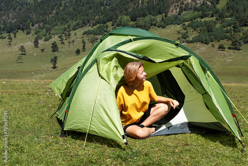Happy child sitting near tourist tent in mountain camping enjoying beautiful summer nature view. A teenage girl traveler enjoying moment, sitting with closed eyes in tourist tent.