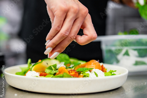 Hand garnishing a vibrant salad with fresh greens, tomatoes, and cheese on a white plate, in a kitchen setting. A moment of meal preparation captured