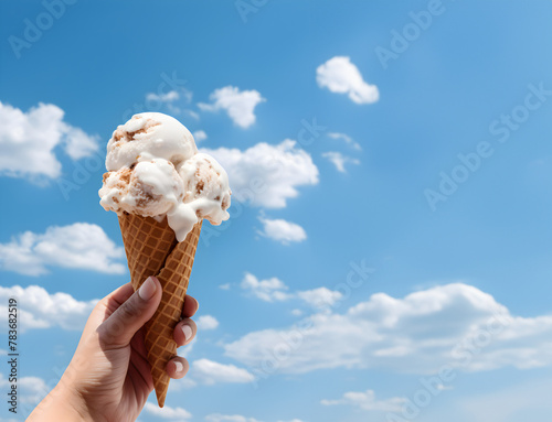 Close up of a childs hand holding a cone with ice creme scoop, blue summer sky background 