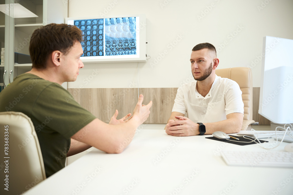 Doctor listening to health complaints of adult man during consultation - obrazy, fototapety, plakaty 