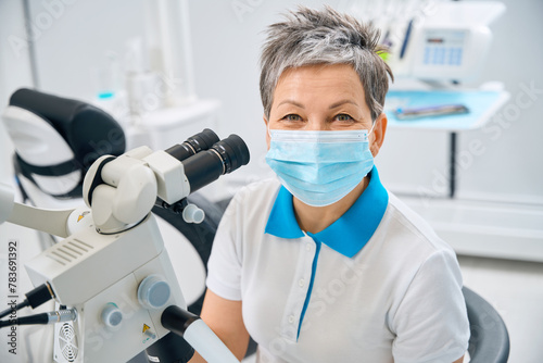 woman oral surgeon in protective face mask sitting near big microscope