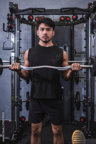A fit asian man in a black tank top does basic standing barbell curls at the gym. Using an EZ curl bar with fixed weight to build biceps.