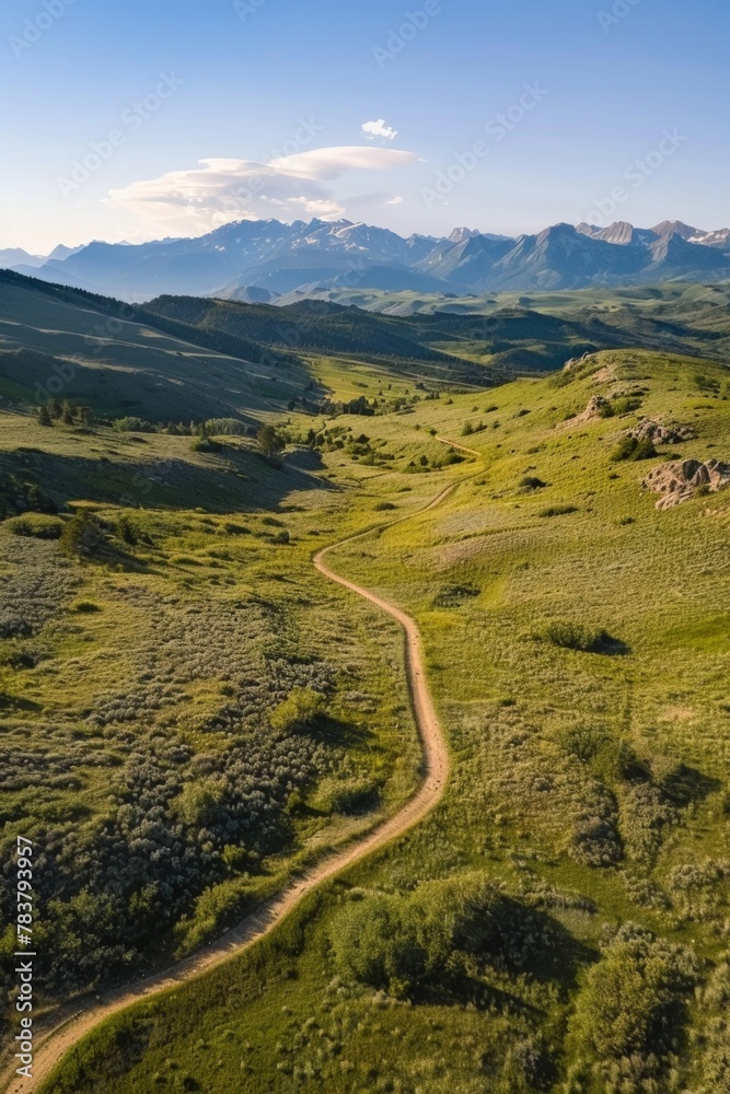 Aerial perspective of a hiking trail winding through a lush green meadow, leading towards a breathtaking mountain range Aerial perspective of a hiking trail winding through a lush green meadow, leading towards a breathtaking mountain range
