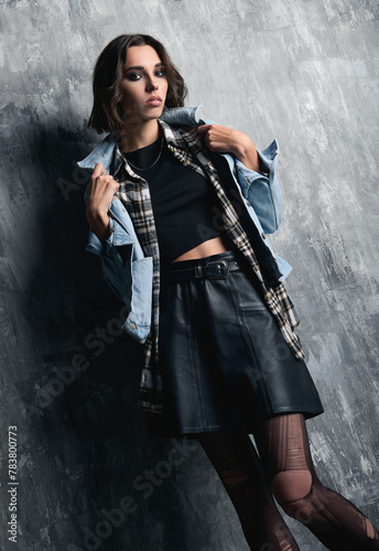 Indoor portrait of pretty grunge (rock) girl standing at wall. Cool informal model, dressed in a jean jacket, checkered shirt, leather skirt and torn tights