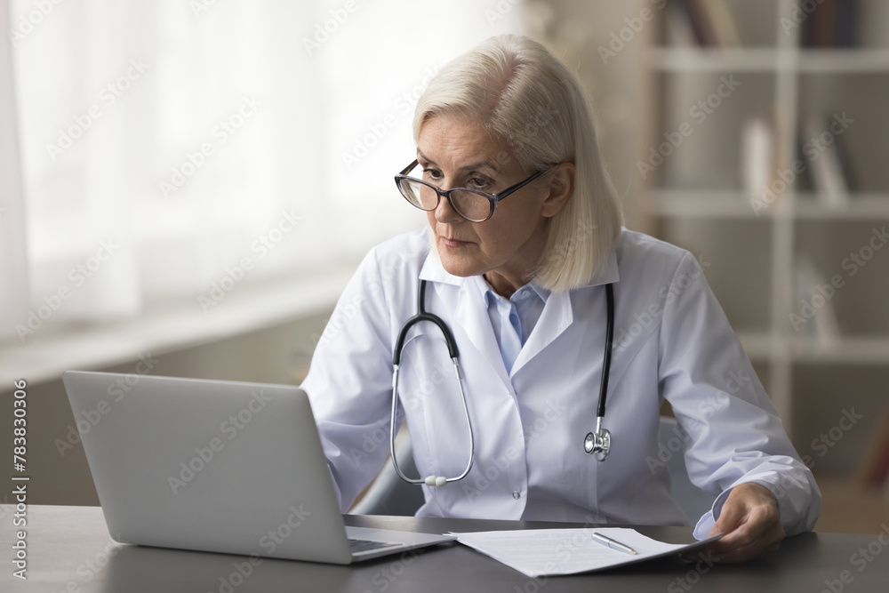 Fototapeta premium Mature woman therapist working on laptop, sit at desk with papers, prepare treatment plan, check patient medical records, make research and clinical guidelines, reviewing data, do administrative tasks