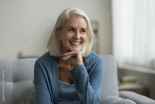 Cheerful attractive aged woman in casual clothes sit on cozy couch in living room, smile, having perfect white teeth, laughing, staring aside, enjoy carefree leisure, comfort and untroubled retirement photo