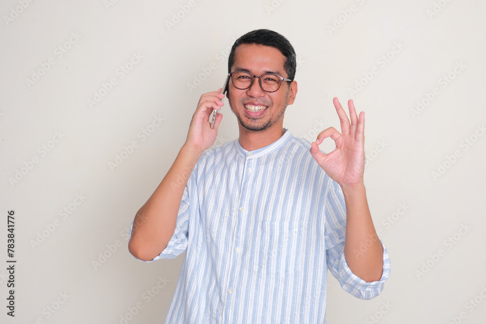 Fototapeta premium Adult Asian man smiling and give OK finger sign while answering a phone call